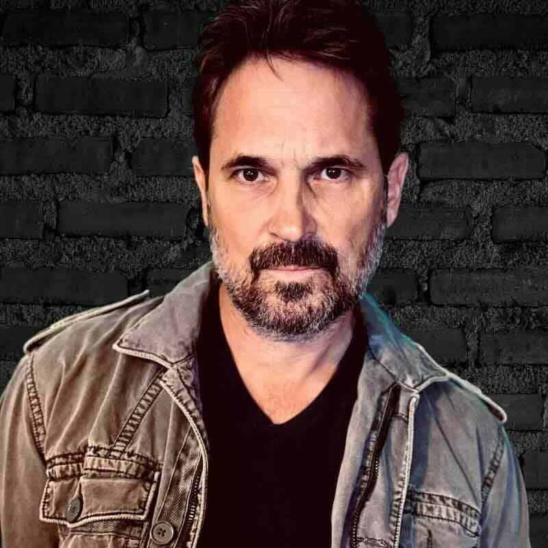 Portrait of a charismatic man with dark hair and beard, wearing a casual jacket, on a black brick background.