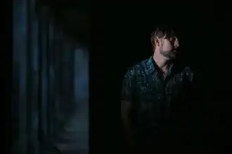 Male figure in patterned shirt standing in darkness with subtle lighting, evoking introspective mood.