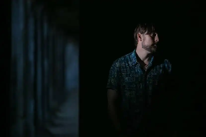 Male figure in patterned shirt standing in darkness with subtle lighting, evoking introspective mood.
