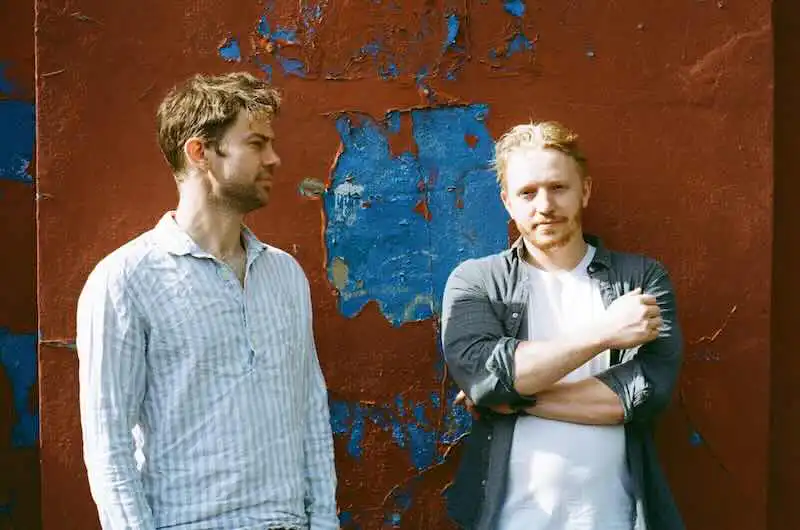 Two men standing outdoors against a weathered blue and red wall, showcasing friendship and connection.