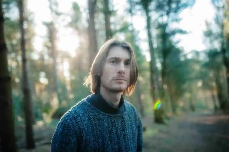Natural outdoor portrait of a young man with long hair in a forest with sunlight filtering through trees.