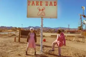 Two individuals standing near a humorous "Fake Dad" sign in a barren desert area. Bright, sunny setting with mountains in the background, highlighting themes of humor and family.