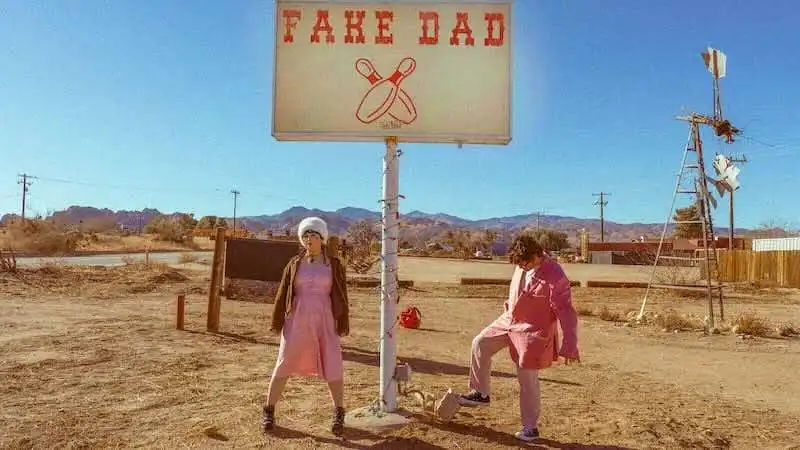 Two individuals standing near a humorous "Fake Dad" sign in a barren desert area. Bright, sunny setting with mountains in the background, highlighting themes of humor and family.