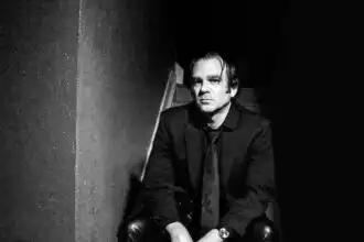 Black and white photo of a stylish man sitting on a chair in a dark setting.