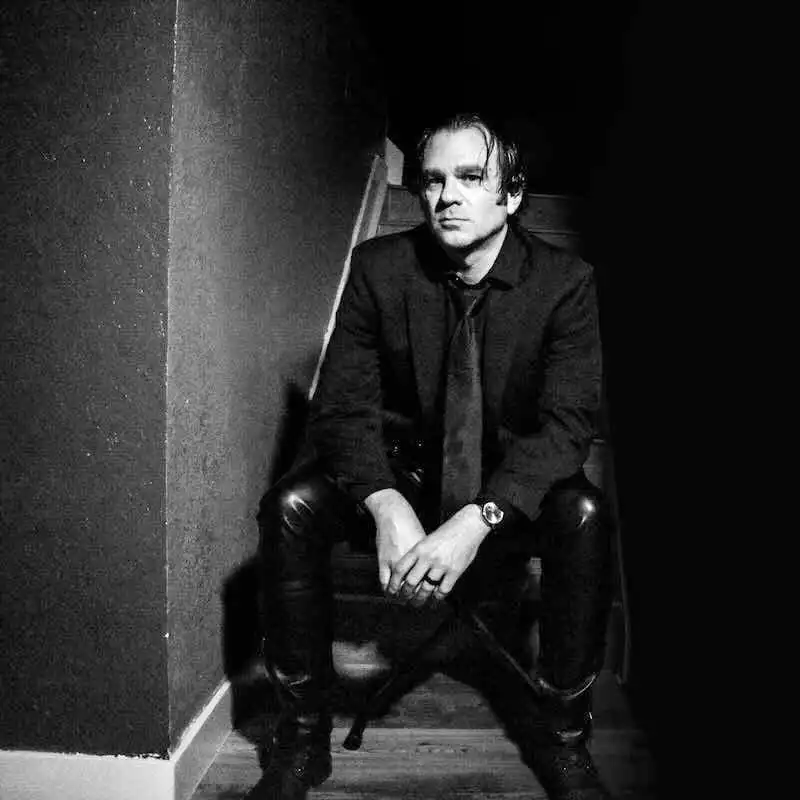 Black and white photo of a stylish man sitting on a chair in a dark setting.