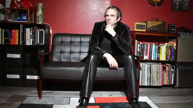 A stylish man in leather pants seated thoughtfully on a black sofa in a cozy, book-filled room.