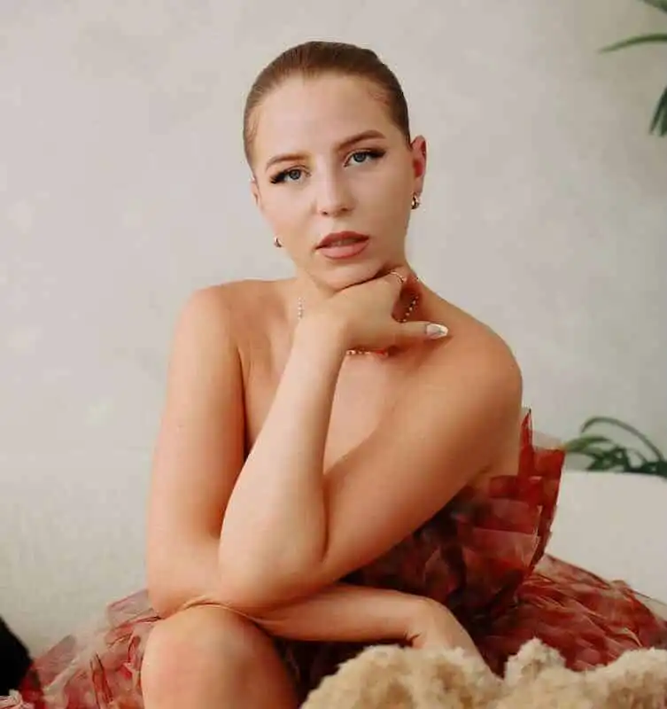 Portrait of a woman with red hair and fair skin, wearing jewelry, and a sheer dress, in a minimalist setting.