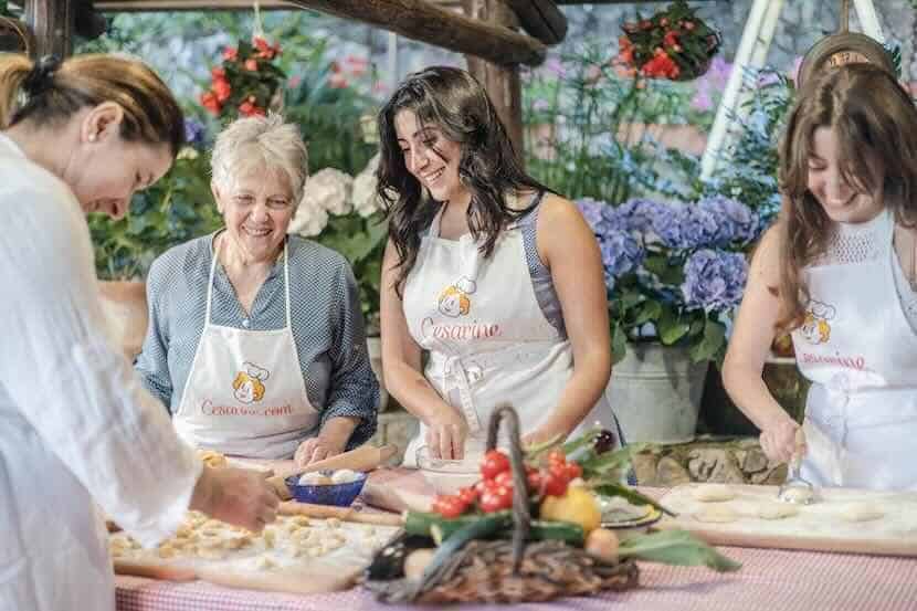 Smiling women participating in a cooking class outdoors wearing aprons, preparing food together with fresh ingredients in a vibrant garden setting.