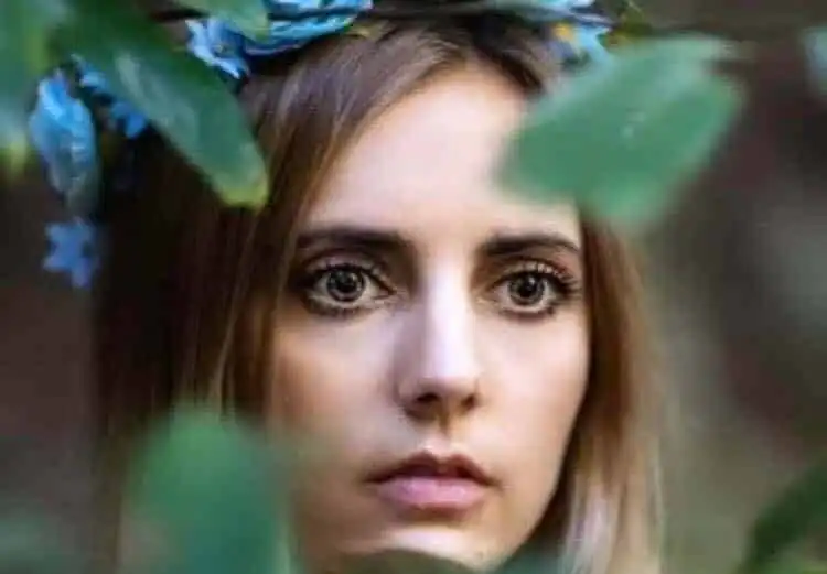 Close-up of a young woman with a floral headband, surrounded by greenery.