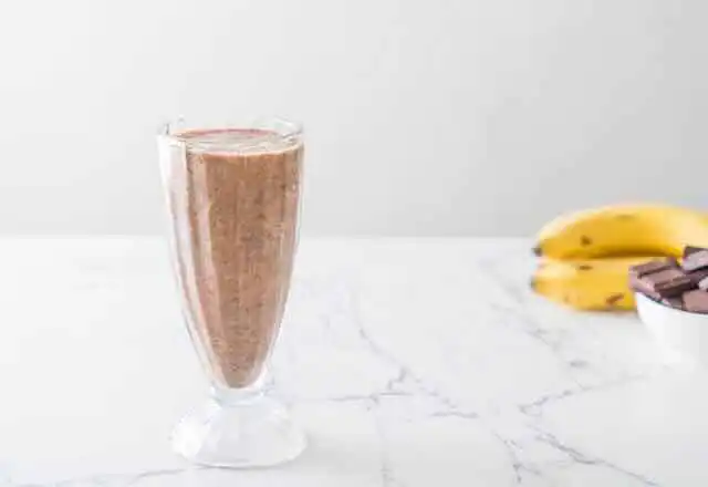 A refreshing banana smoothie topped with cocoa powder, served in a tall clear glass on a white marble surface.