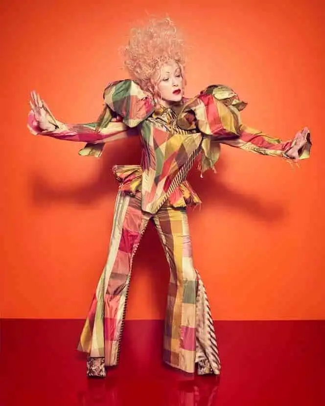 An avant-garde fashion runway model wearing colorful, bold patterned clothing against a vivid orange background.