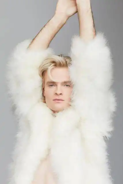 A stylish man with platinum blonde hair wears a luxurious white fur coat, showcasing fashion and confidence.