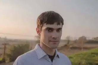 Young man standing in field at sunset, wearing a checkered shirt, peaceful expression.