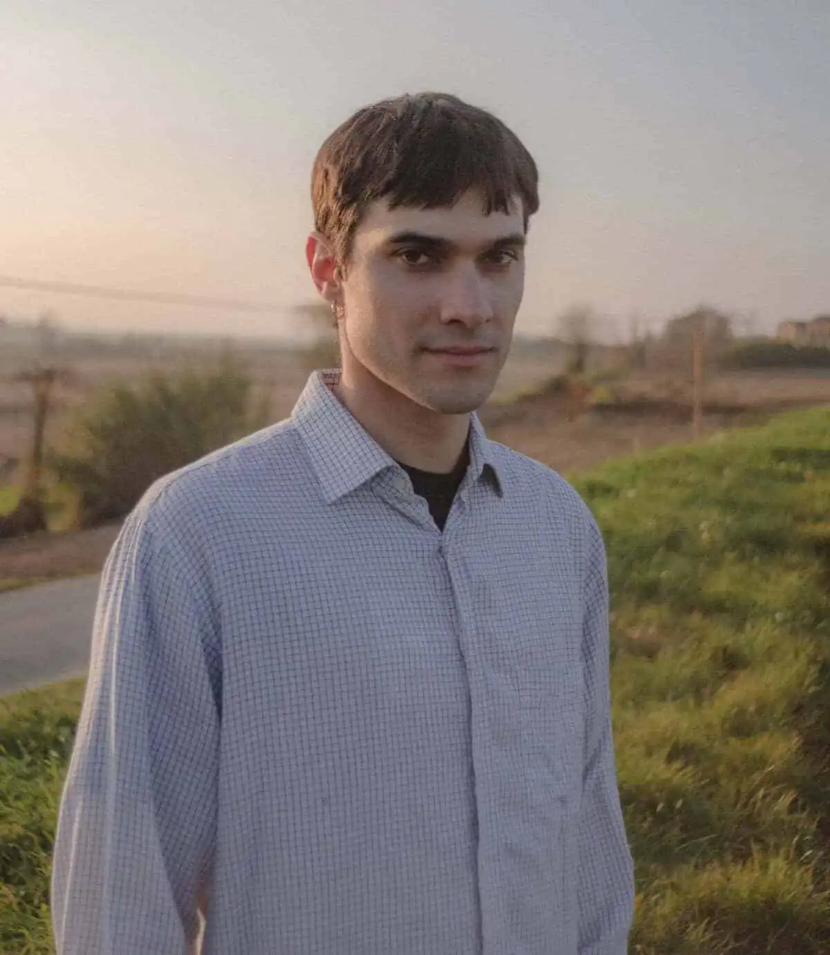 Young man standing in field at sunset, wearing a checkered shirt, peaceful expression.