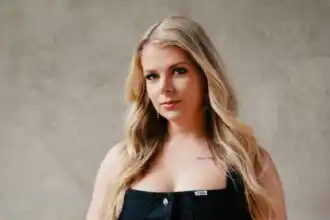 Blonde woman with wavy hair and subtle makeup, wearing a black top, posing confidently.