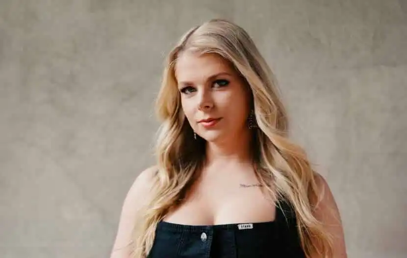 Blonde woman with wavy hair and subtle makeup, wearing a black top, posing confidently.