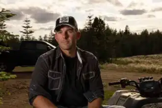 Smiling man sitting outdoors with trees and sunset sky in background.
