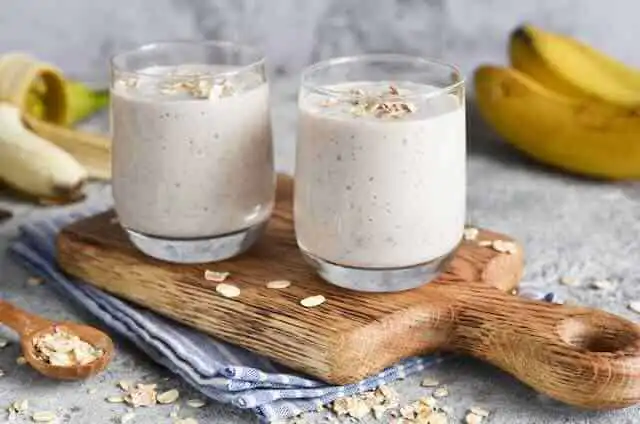 Creamy banana oatmeal smoothie in two glasses, with bananas and oats in the background.