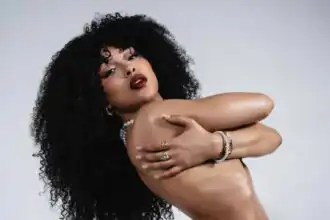 Black woman with curly hair and bold makeup, topless, with red roses, jewelry, and confident pose.