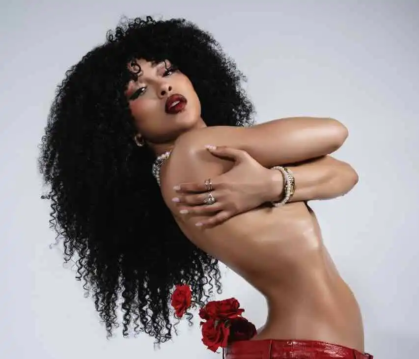 Black woman with curly hair and bold makeup, topless, with red roses, jewelry, and confident pose.