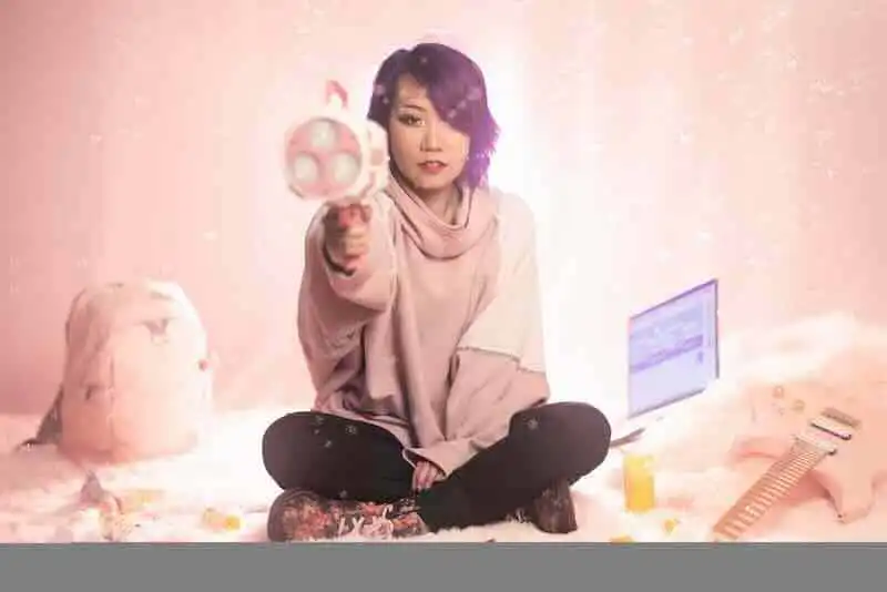 A confident woman sitting on the floor, holding a toy gun, with a laptop and backpack in a cozy, playful setting.