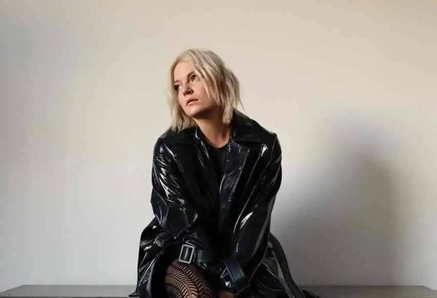 Woman in black leather jacket poses thoughtfully against plain background.