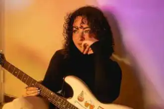 Close-up of woman with curly hair holding an electric guitar, relaxed and thoughtful in a warmly-lit room.