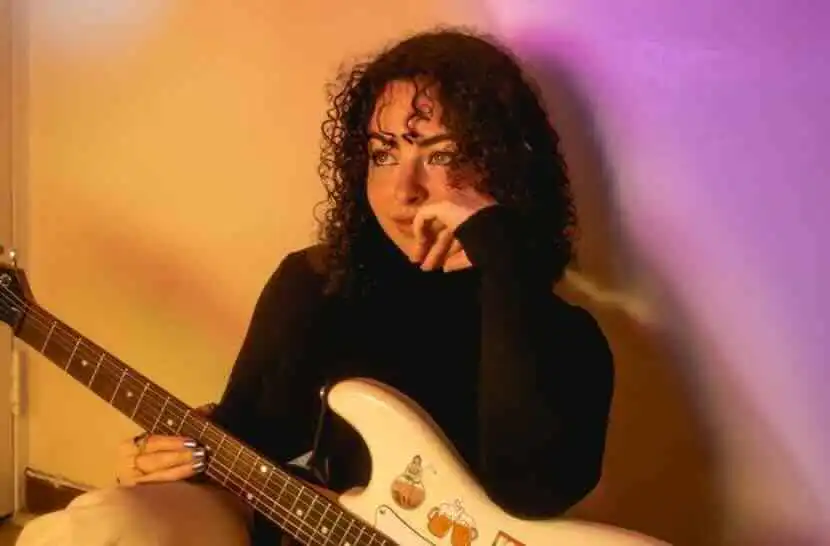 Close-up of woman with curly hair holding an electric guitar, relaxed and thoughtful in a warmly-lit room.