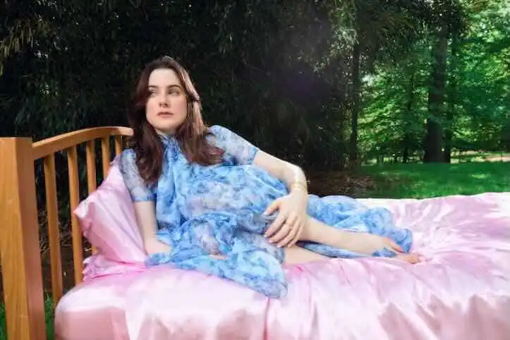 Woman relaxing outdoors on a pink bed with a blue dress, lush green trees background.
