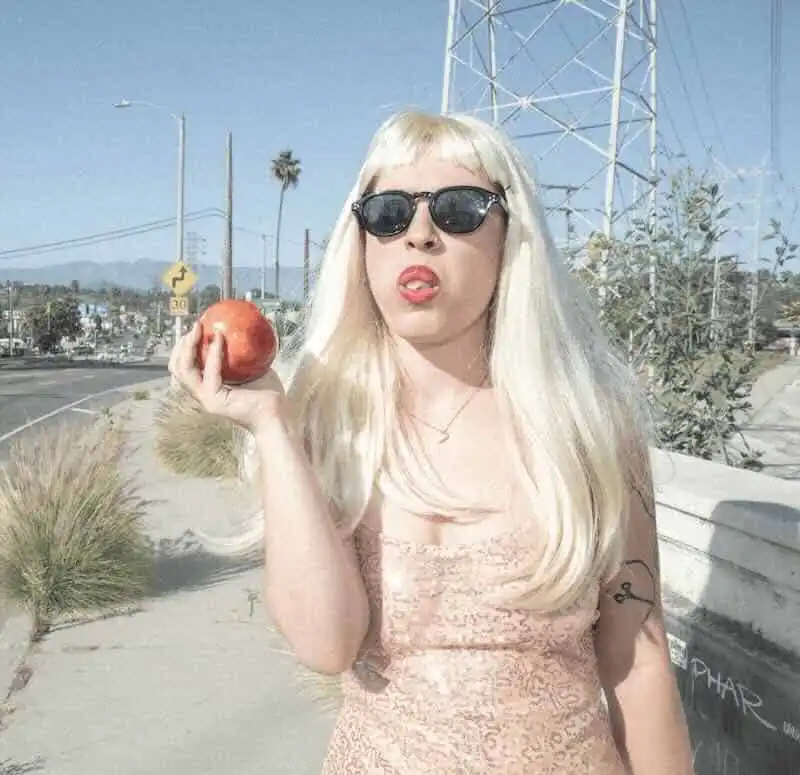 Woman with blonde hair and sunglasses holding an apple outdoors.