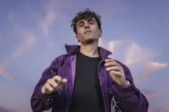 A confident man with curly hair dressed in a purple jacket and black shirt, standing outdoors against a dreamy sky, showcasing pride and self-expression.
