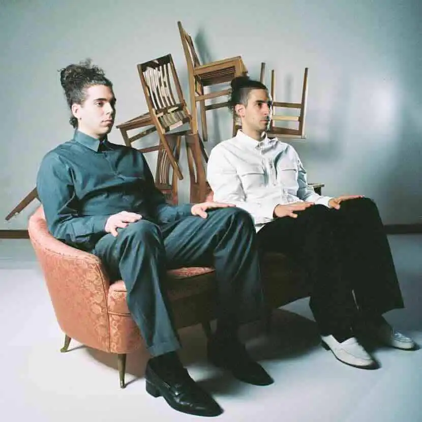 Men sitting on a pink velvet couch with stacked chairs in background.