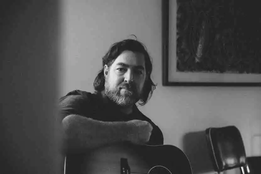 A black and white image of a gay man with a beard playing guitar at home, showcasing his artistic and creative personality.