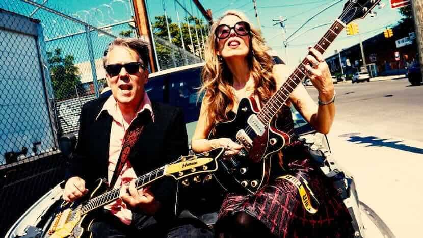 Vibrant image of a gay male and female musician playing guitars outdoors in an urban setting, showcasing LGBTQ+ pride and musical talent.