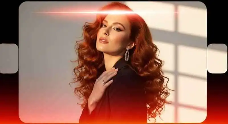Glamorous red-haired woman with curly hair, elegant makeup, and stylish jewelry poses confidently.