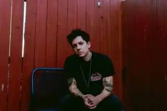 Portrait of a young man with dark hair and tattoos sitting against a red wooden fence.