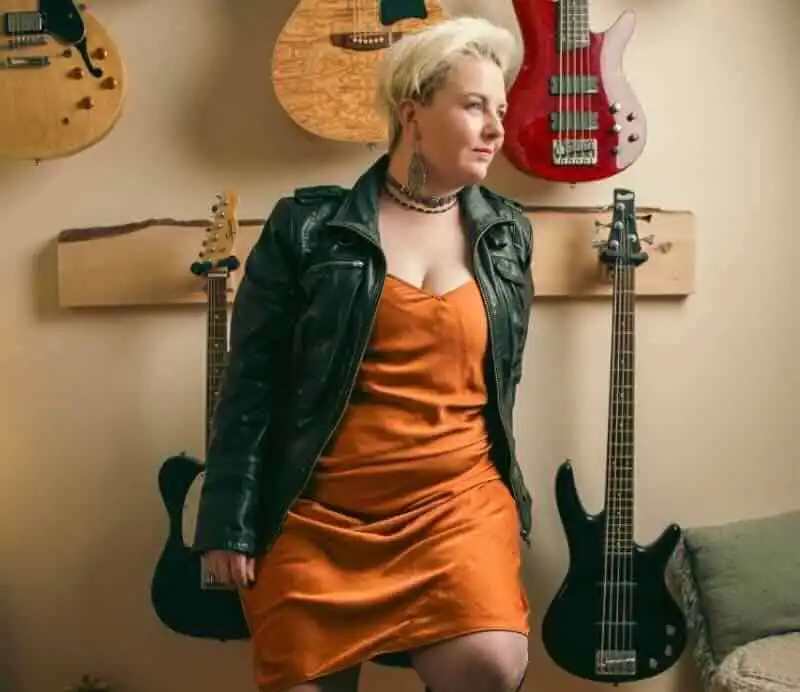 Young woman with short blonde hair and edgy style standing among guitars.