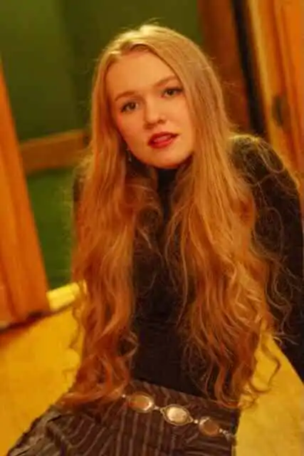 Young woman with long red hair, wearing a black top, sitting indoors at Divine Magazine.