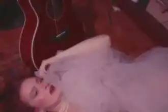 Elegant woman with vintage pearl jewelry lying on a plush pink bed, guitar in background, stylish scene.