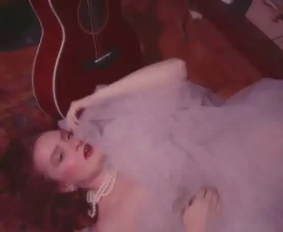 Elegant woman with vintage pearl jewelry lying on a plush pink bed, guitar in background, stylish scene.