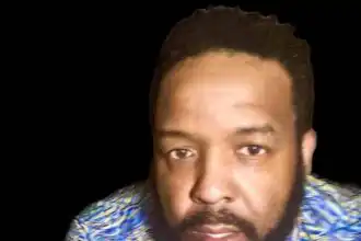 Close-up of a confident, stylish man with a beard wearing a colorful shirt against a black background.