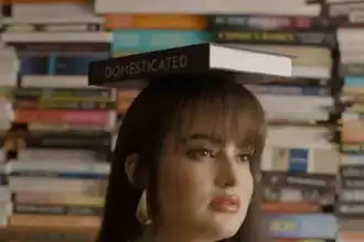 Close-up of a woman with a book on her head, surrounded by a stacked book background, conveying literary and intellectual themes.