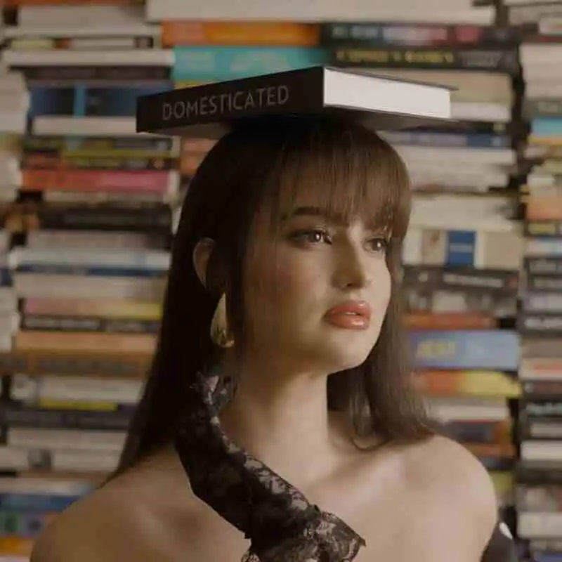 Close-up of a woman with a book on her head, surrounded by a stacked book background, conveying literary and intellectual themes.