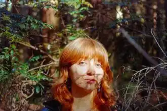 Red-haired woman outdoors among foliage and branches, natural light and shadows.