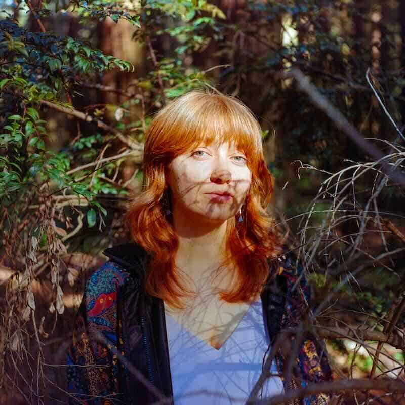 Red-haired woman outdoors among foliage and branches, natural light and shadows.