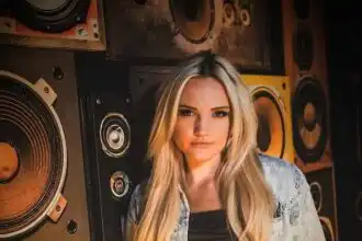A young woman with long blonde hair stands in front of speakers in a music studio, wearing a denim jacket and black top, showcasing a confident attitude.