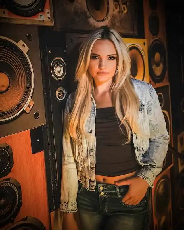 A young woman with long blonde hair stands in front of speakers in a music studio, wearing a denim jacket and black top, showcasing a confident attitude.