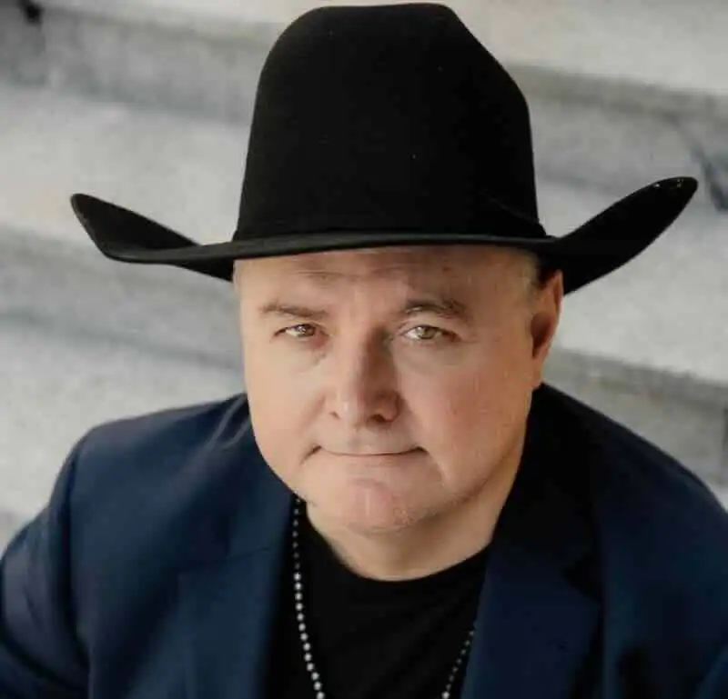 Close-up of a confident man wearing a black cowboy hat and dark blazer, expressing strength and style.
