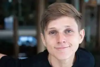 Young man smiling, casual attire, by a scenic outdoor background.