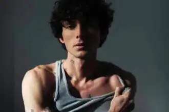 A striking portrait of a tattooed man with curly hair, wearing a tank top against a dark background.